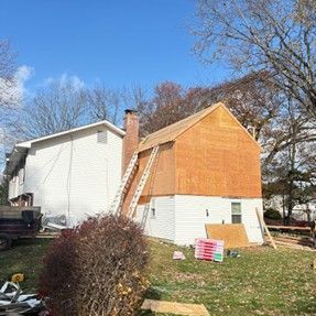 House under construction with plywood siding, ladder against chimney, blue sky, and yard.