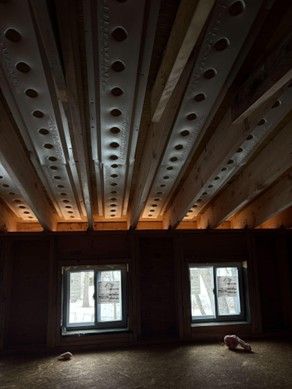 Interior view of a building under construction, showing wooden beams with drilled holes and two windows.