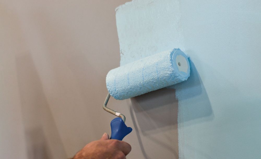 Person painting a wall light blue with a paint roller, applying the paint.