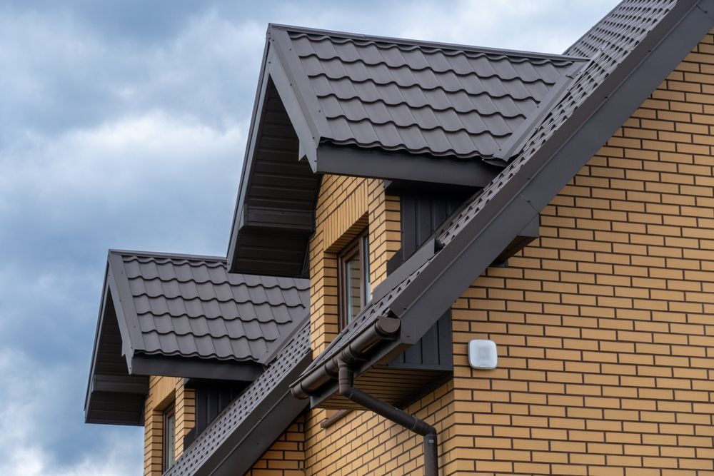 Brick building with dark gray roof and gutters against a cloudy sky.