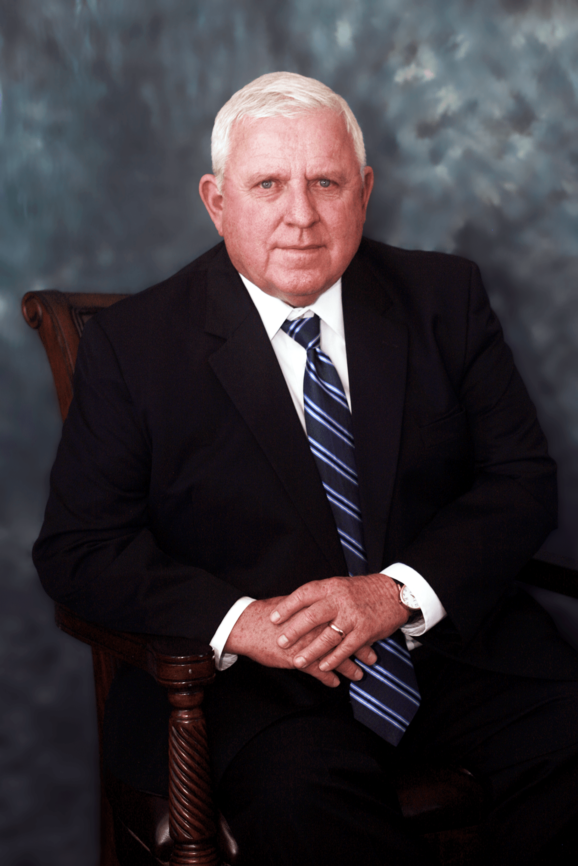 Man in dark suit, seated, hands clasped. White hair, blue striped tie. Dark background.
