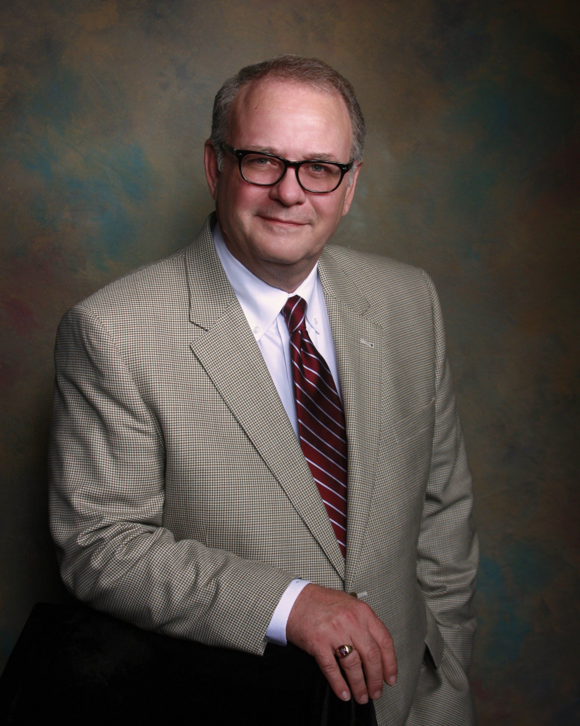 Man in a light-colored suit and glasses smiles, leaning against a dark object.