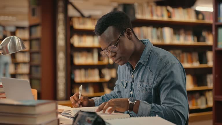 A person wearing glasses and a denim shirt sits at a library desk, focused on writing in an open notebook near a laptop.