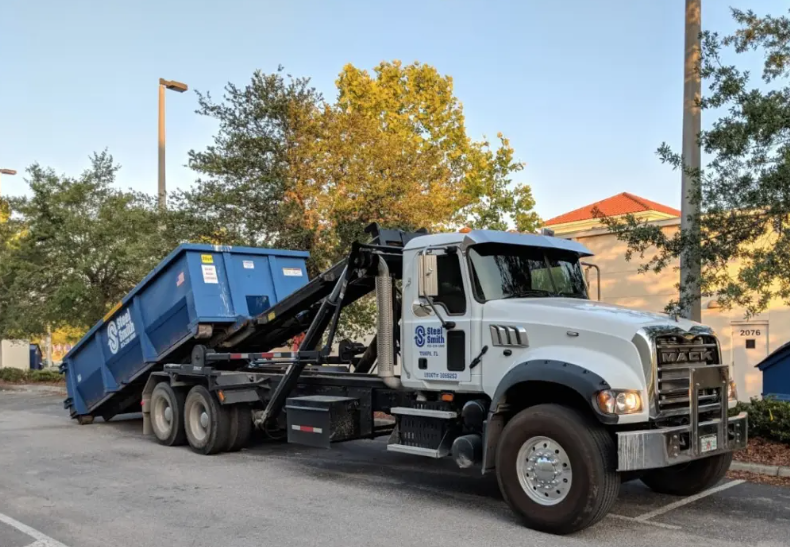 a dump truck is carrying a blue dumpster in a parking lot .