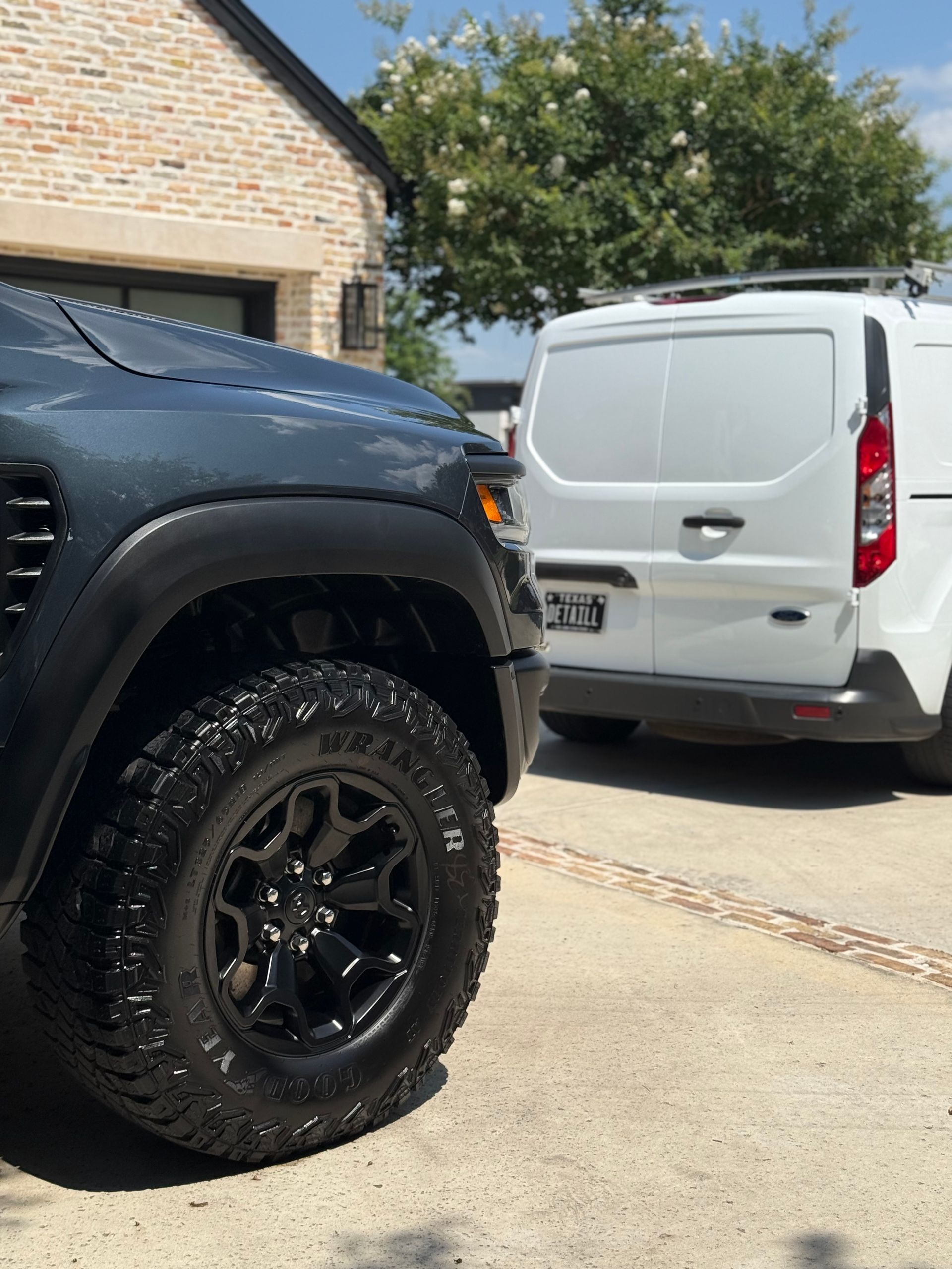 Dark truck with a large tire parked beside a white cargo van, in a driveway.