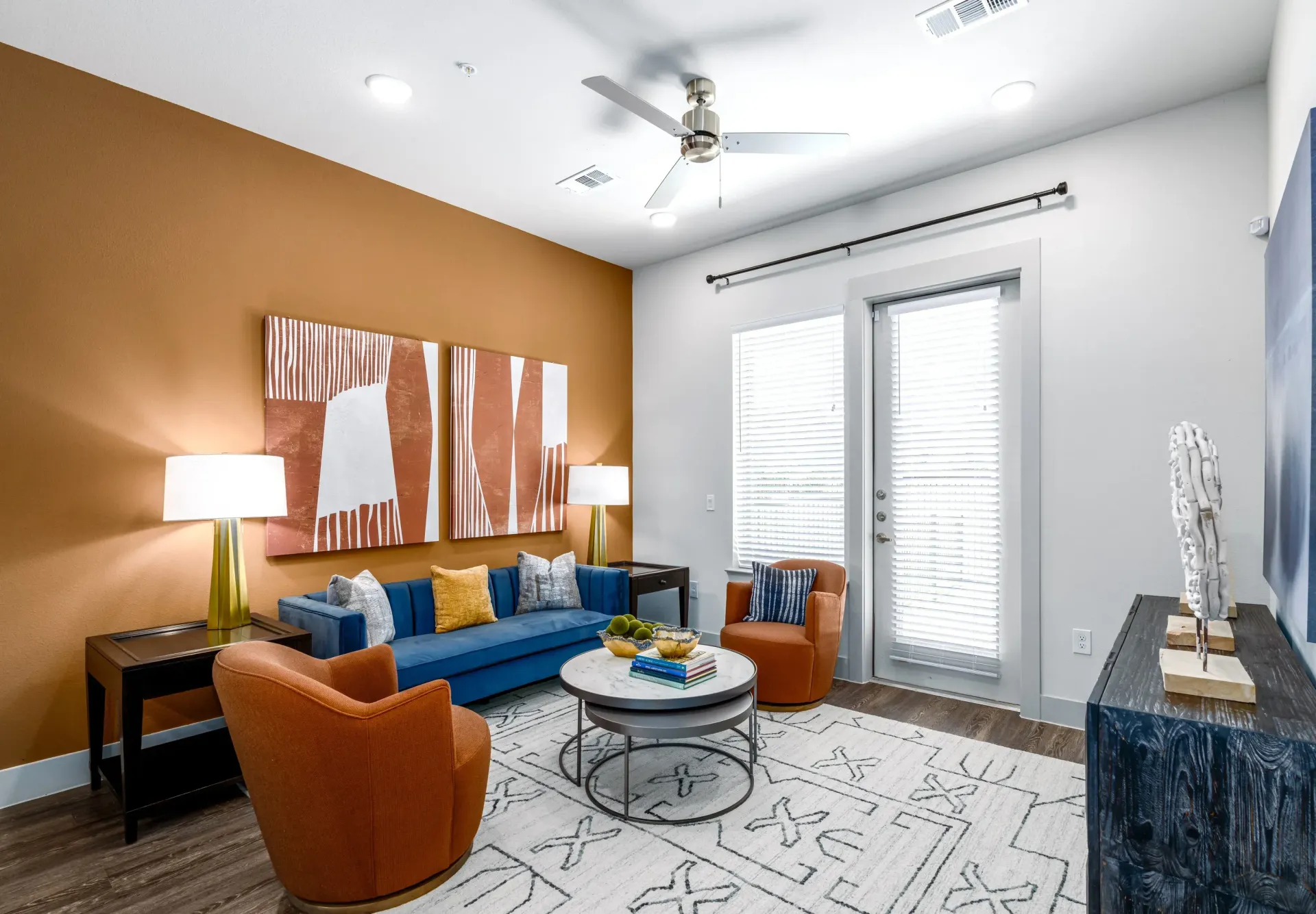 Living room in an apartment with orange accent wall, blue sofa, orange chairs, and modern decor.