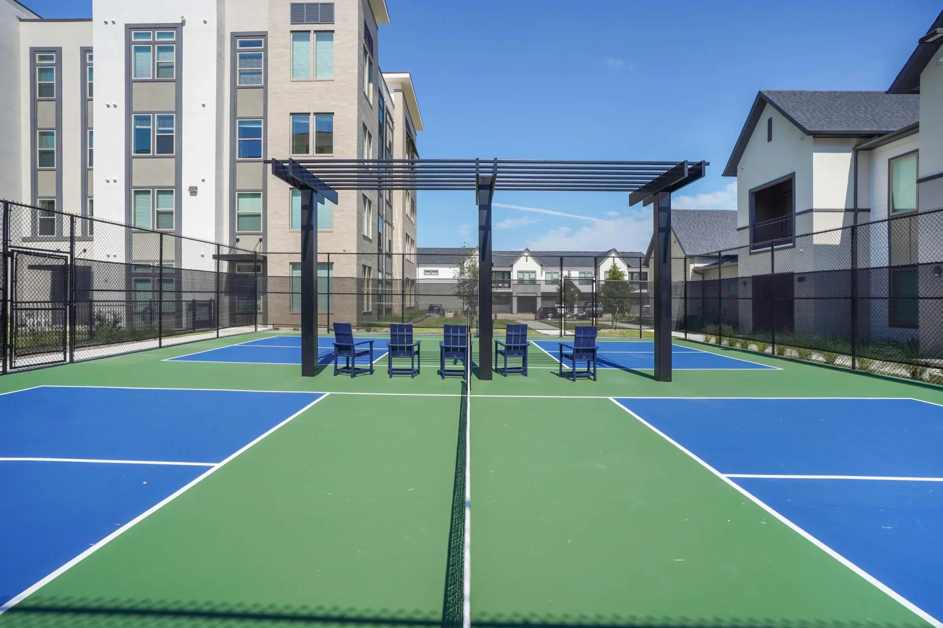 Outdoor community tennis/pickleball court with blue and green surface, seating, and surrounding apartments.
