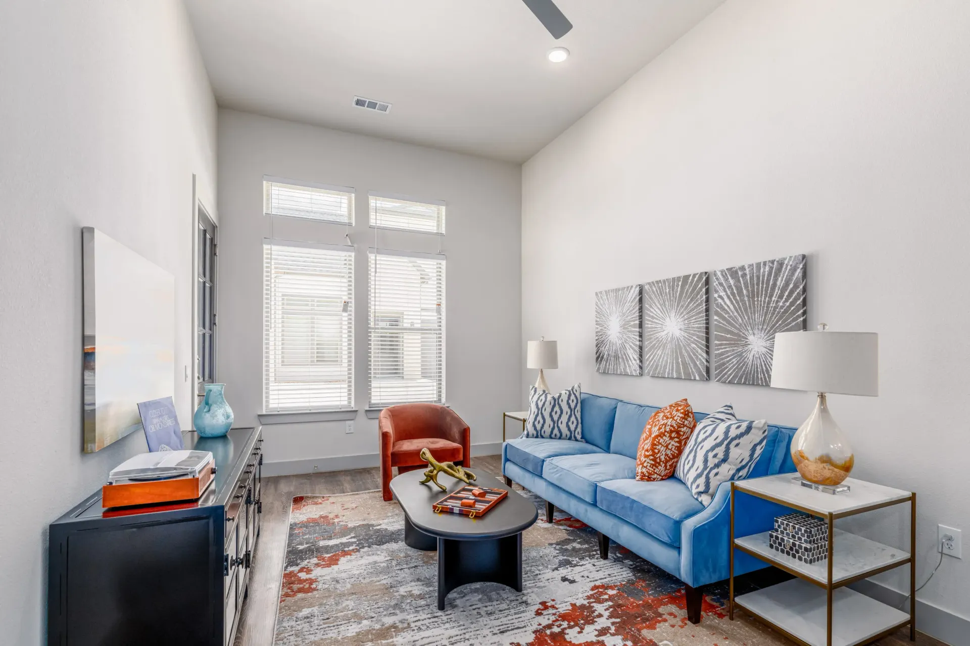 Living room in an apartment with a blue sofa, orange chair, coffee table, and wall art near large windows.