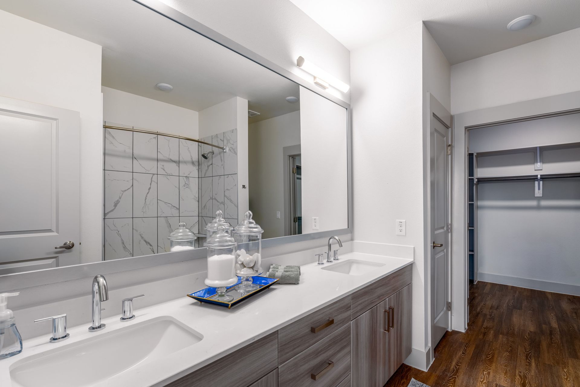 Bright bathroom vanity with double sink, large mirror, and open closet with hardwood floor