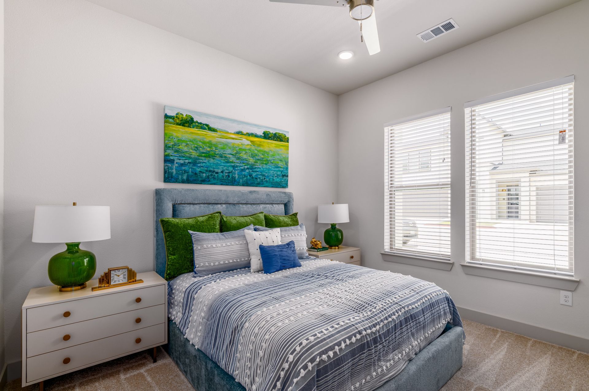 Bright bedroom with a blue patterned bedspread, green accents, and large windows with blinds