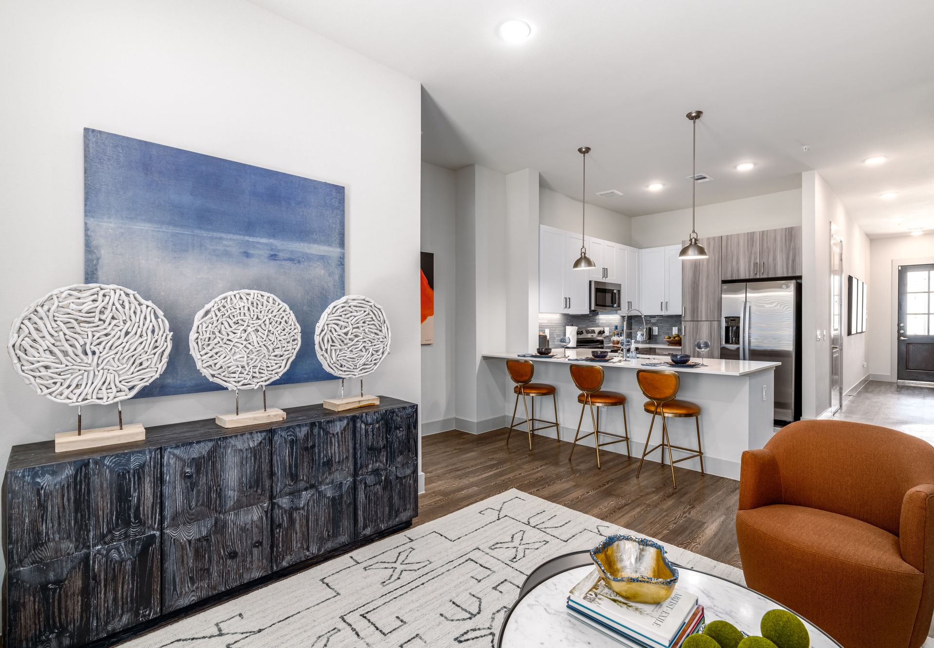 Modern open-plan living room and kitchen with white island, orange stools, brown chair, and blue wall art