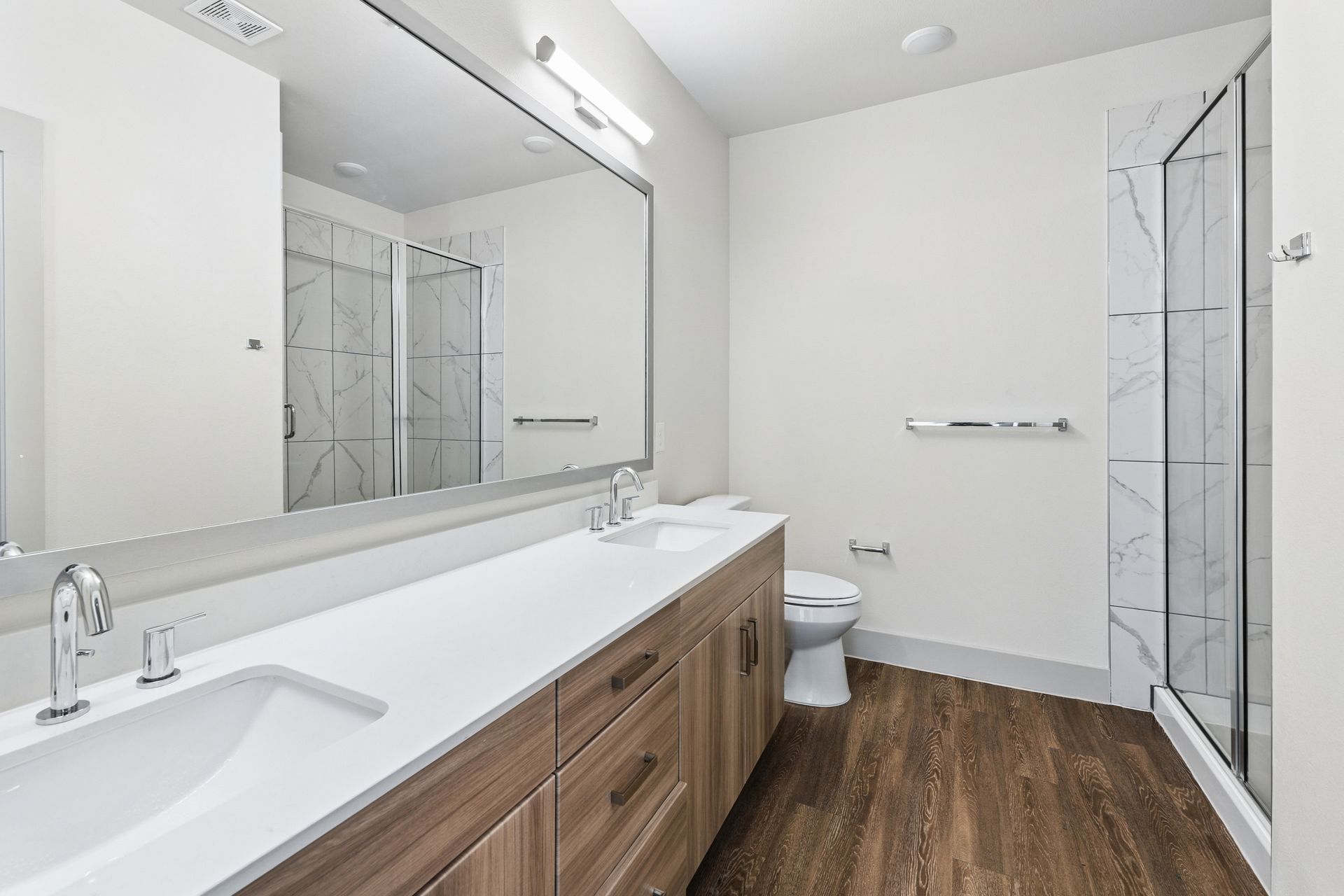 Modern white bathroom with wood vanity, large mirror, glass shower, and toilet