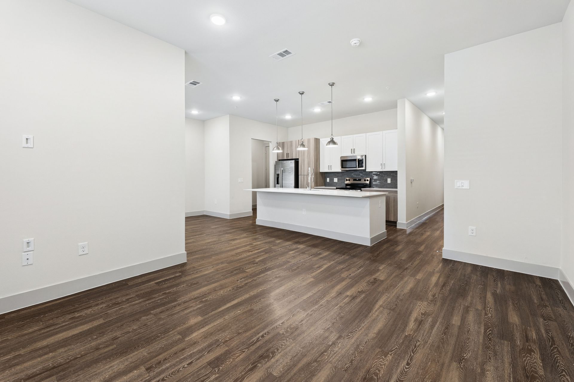 Empty modern kitchen and living area with white walls, dark wood floors, and a white island.