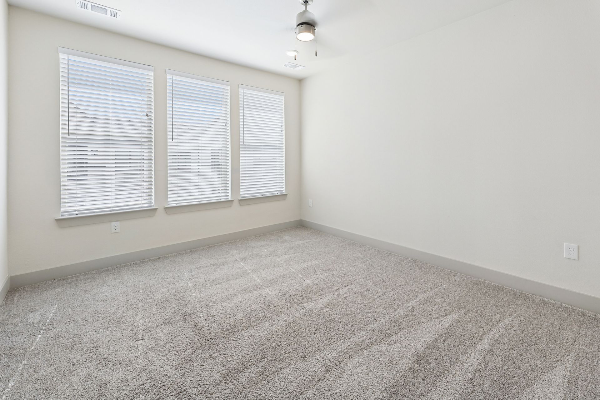Empty white bedroom with carpeted floor, three windows with blinds, and a ceiling light fixture