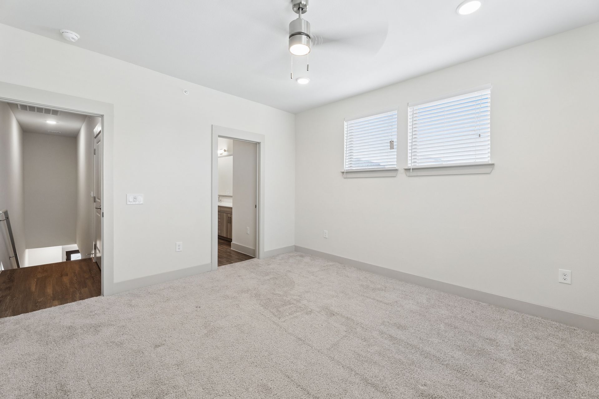 Empty gray-carpeted bedroom with white walls, ceiling fan, mirrored closet doors, and two windows