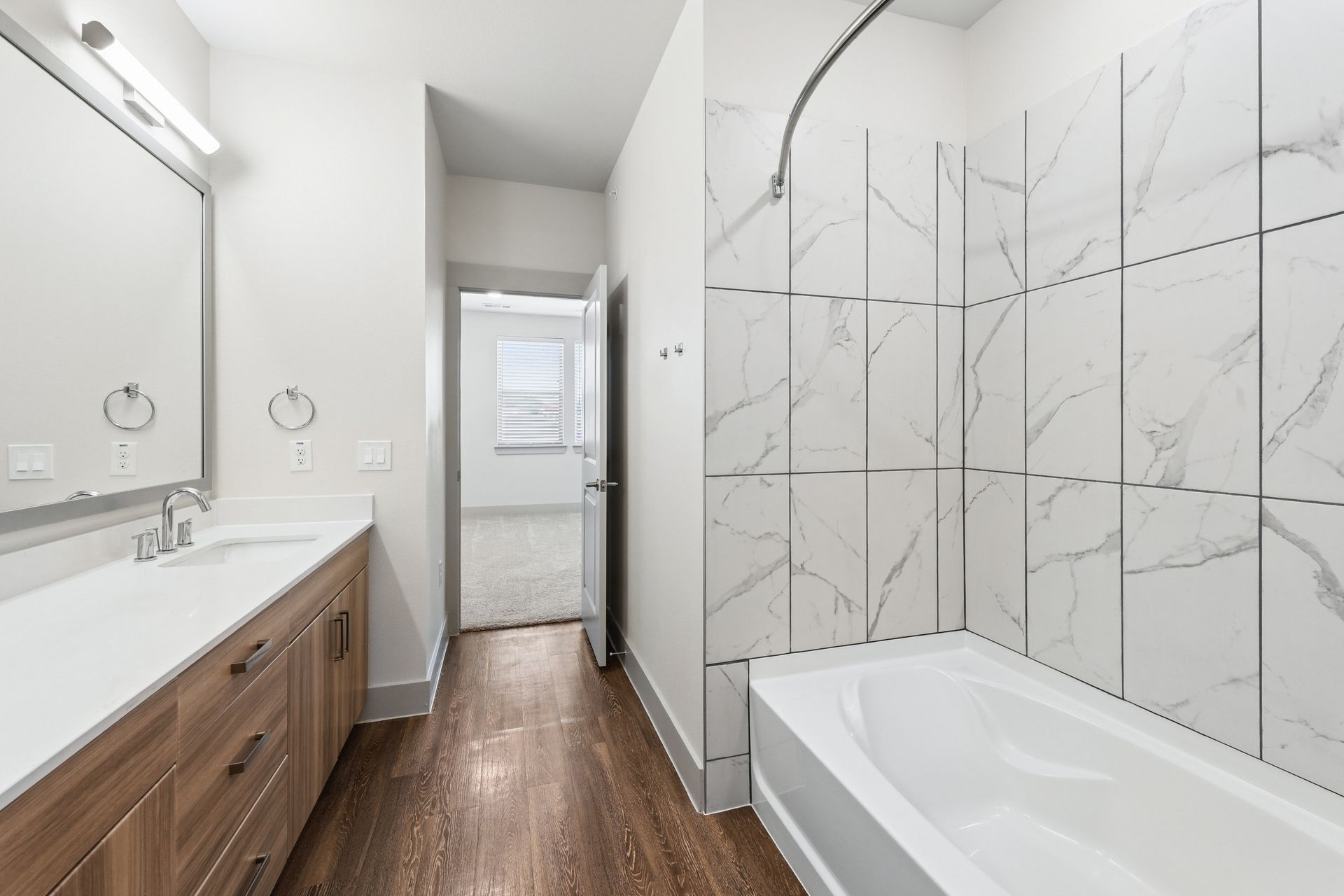 Bright bathroom with wood vanity, large mirror, and white tiled tub/shower along a narrow hallway.