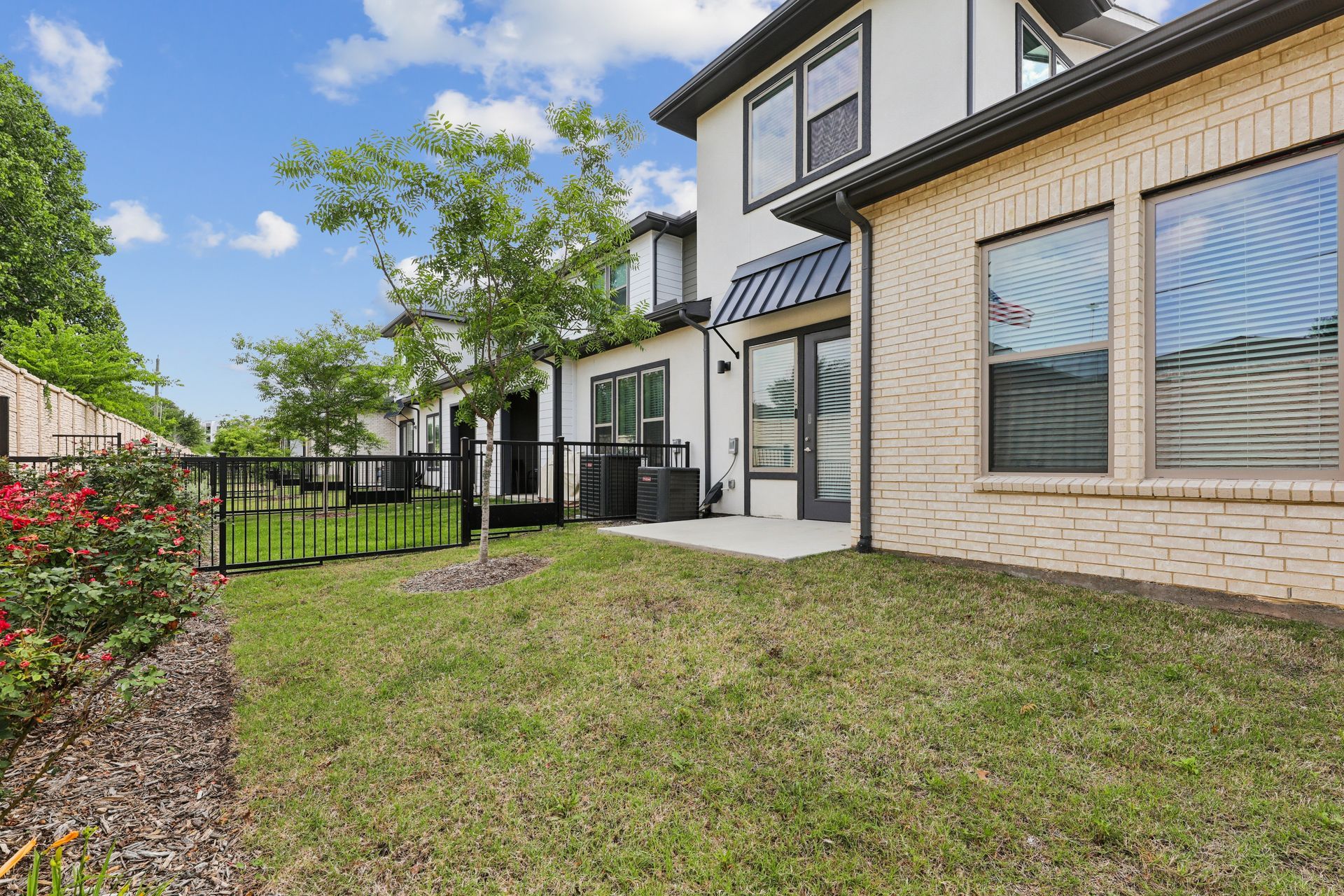 Townhouse backyard patio with grass, black fence, and large windows under a blue sky
