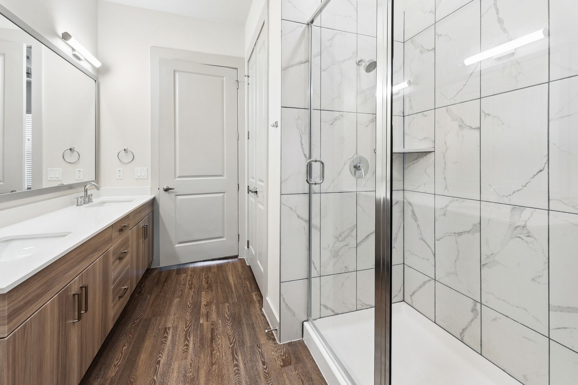 Bright modern bathroom with double vanity, wood cabinets, glass shower, and white marble tile.