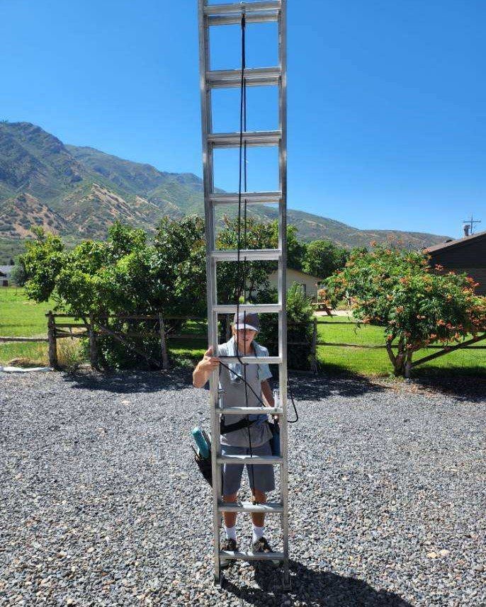 Man standing on ladder; holding equipment; outdoors, gravel driveway, mountain background.