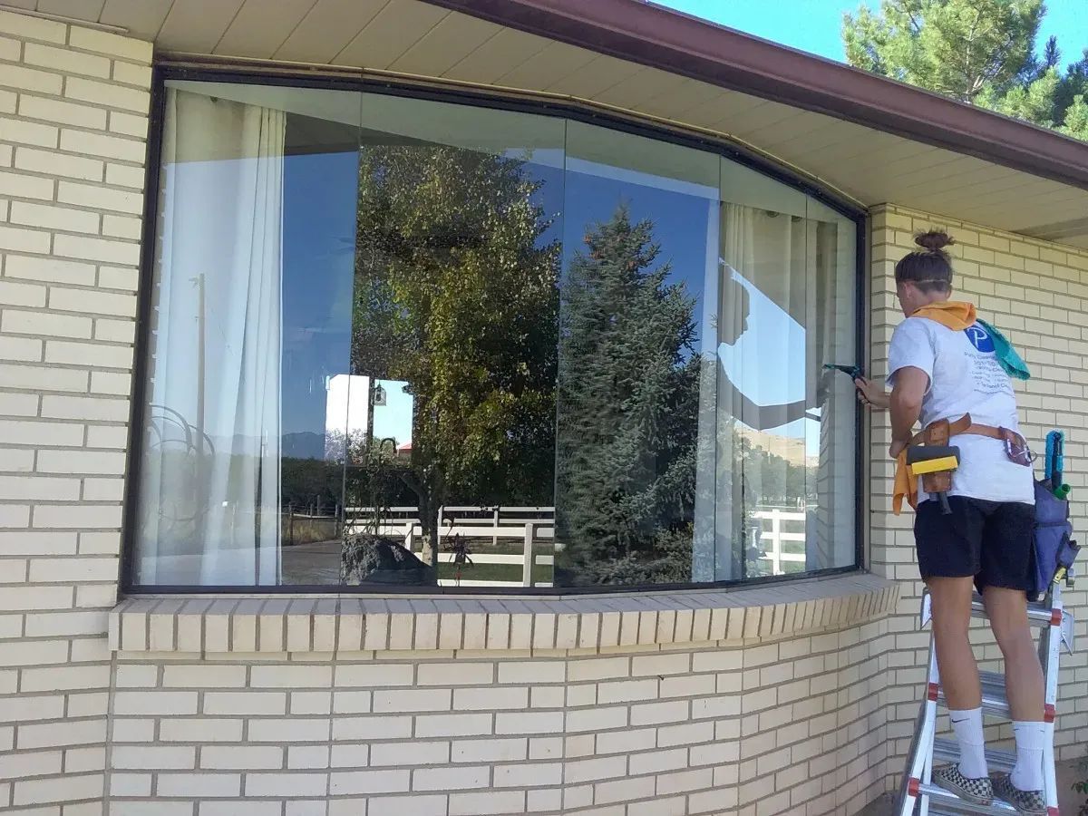 Person on ladder washing curved window on brick building. Trees reflect in the glass.