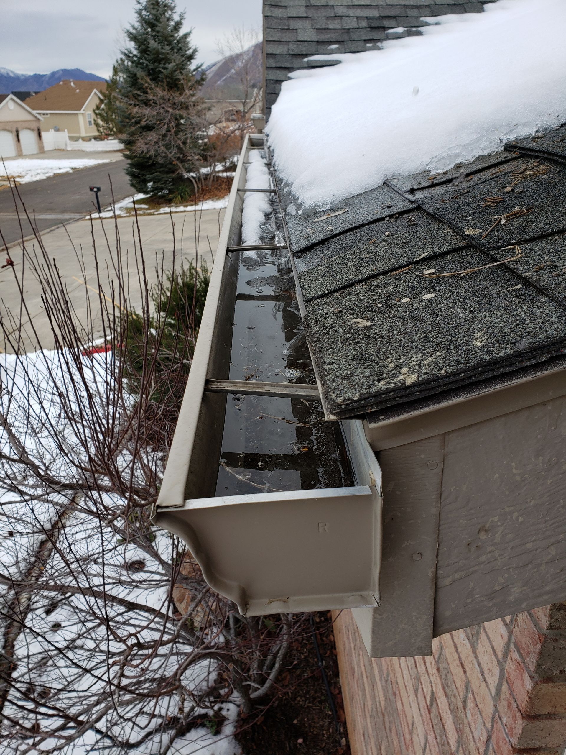 Gutter filled with water and debris; snow on roof. Beige gutters on a brick home in winter setting.