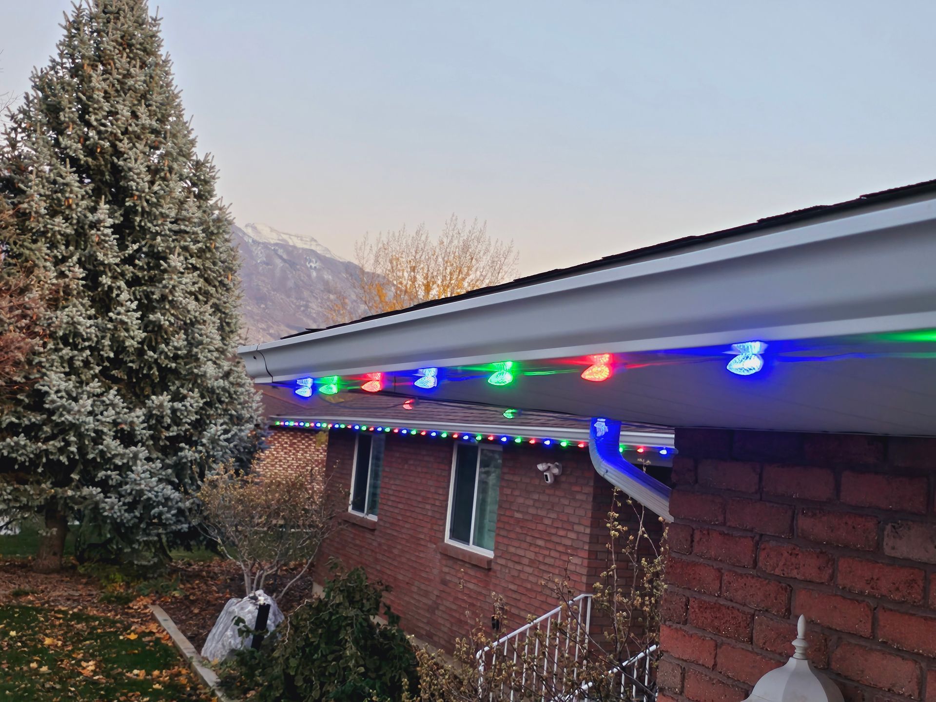 Christmas lights line the gutter of a brick house, lit with blue, green, and red lights.