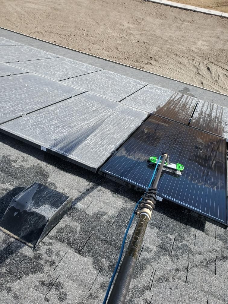 Solar panels being cleaned on a roof with a long-handled brush and water, outdoors.