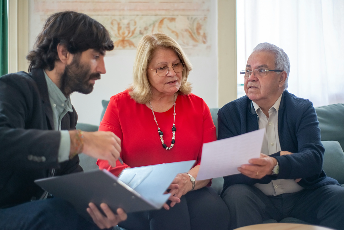Three coworkers discussing documents on a couch in a bright office, one in a red sweater.