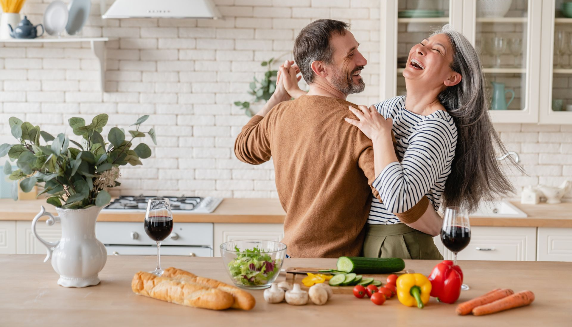 Couple dancing and laughing in a bright kitchen, with a wooden countertop and fresh vegetables.