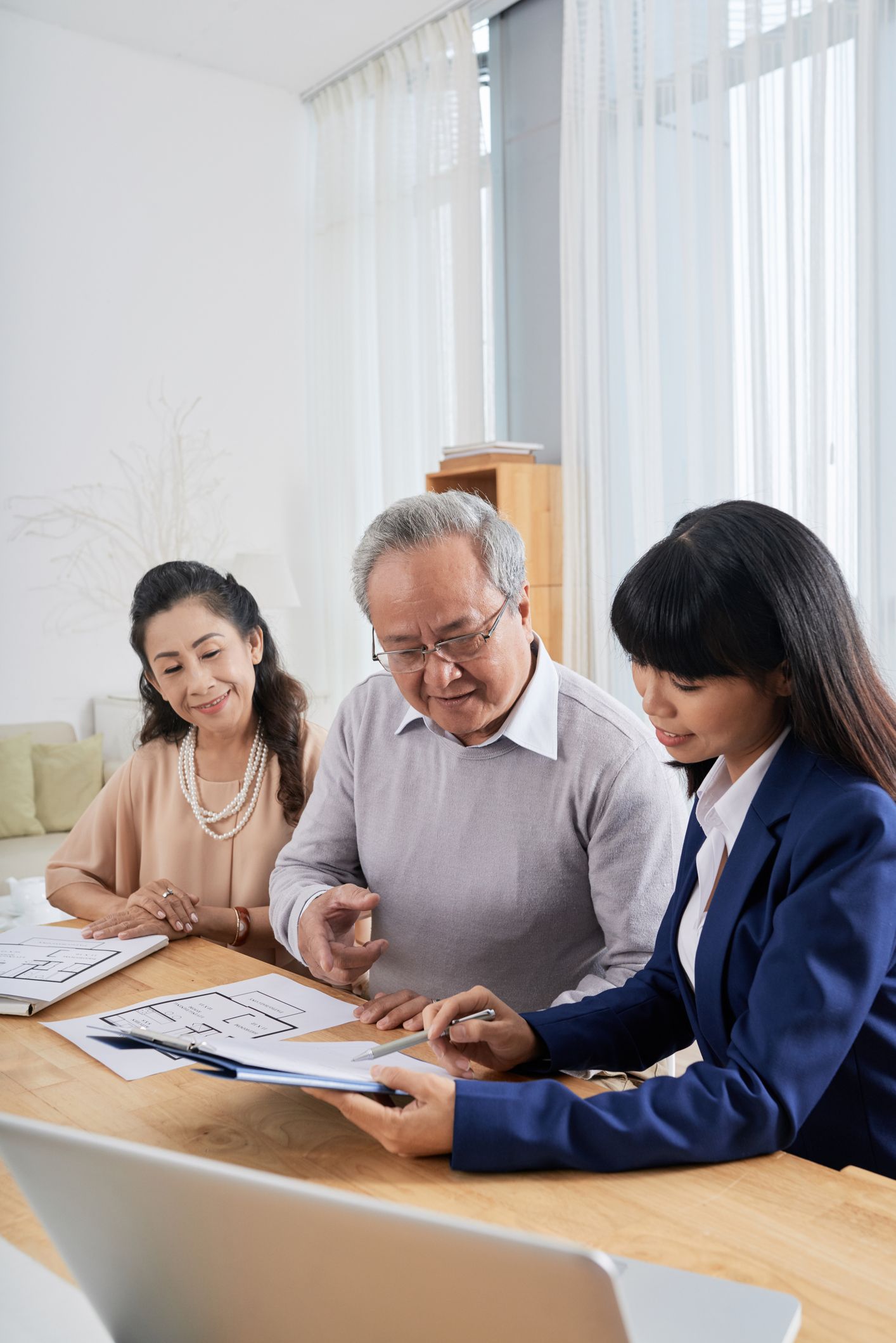 Woman in suit reviews paperwork with a couple at a table. Sunlight streams in from a window.
