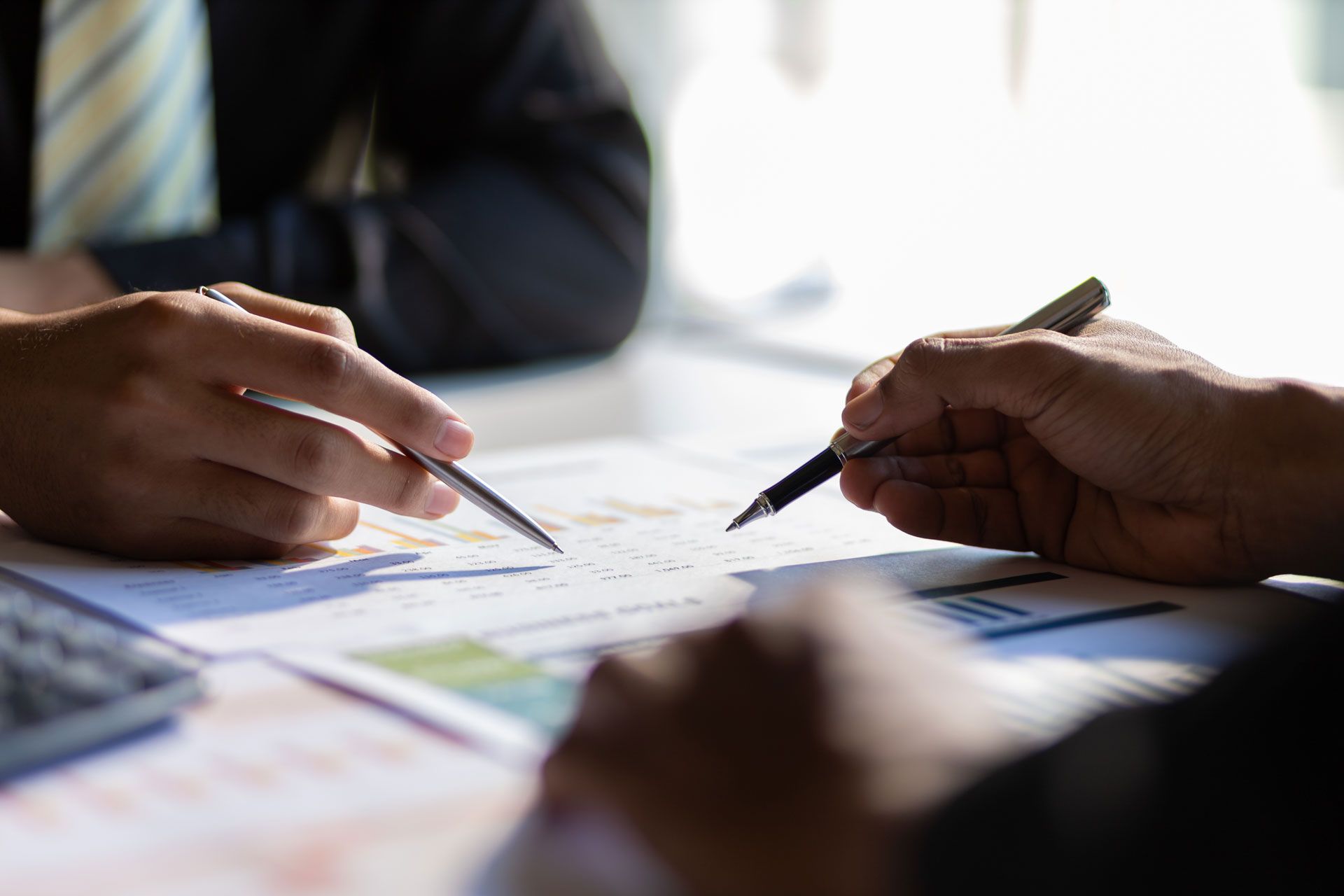 Two people reviewing paperwork with pens, focusing on charts and graphs.