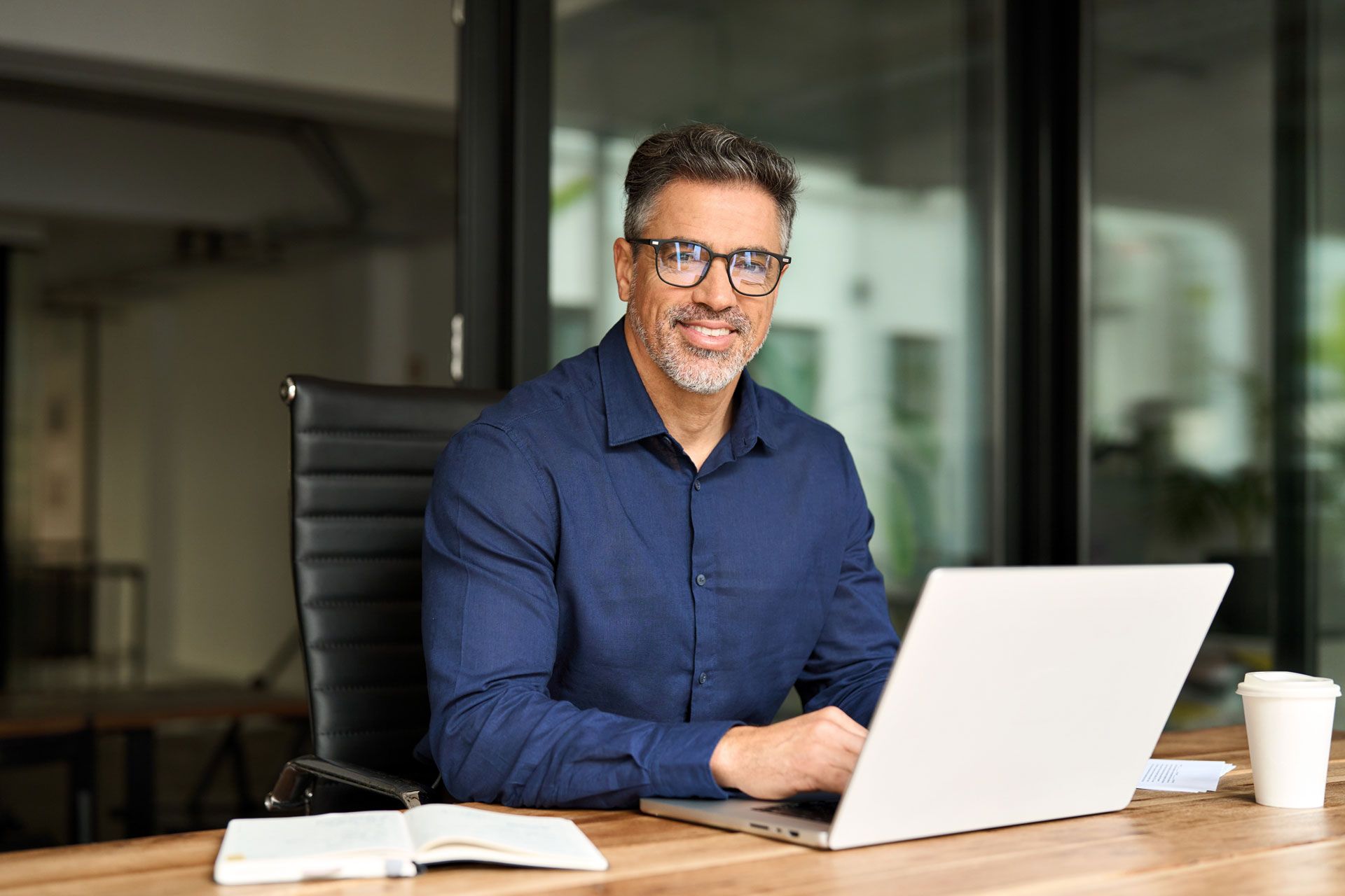 Man in blue shirt, glasses, smiling at laptop in office setting.