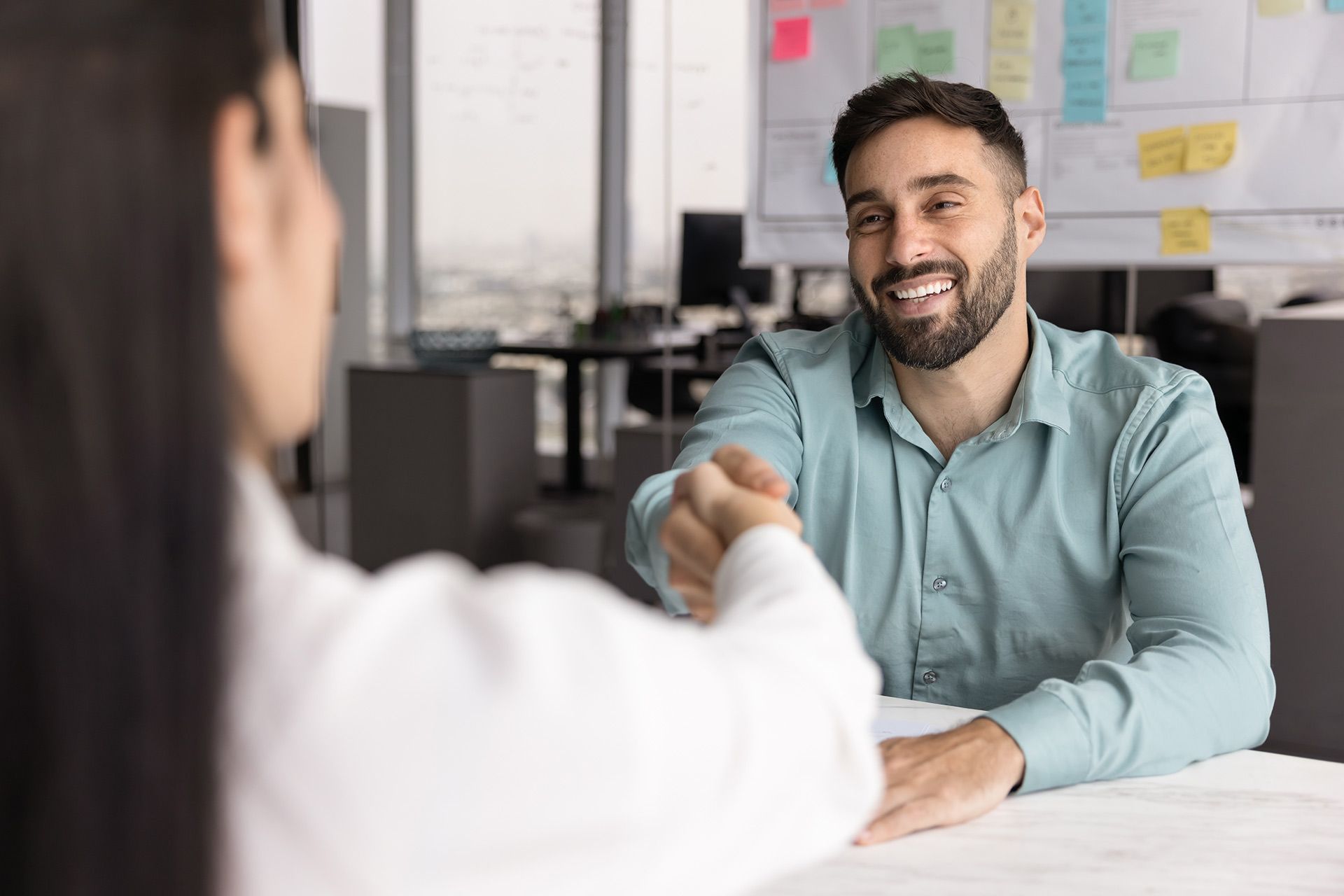 Man in blue shirt smiling, shaking hands with person at a desk in an office setting.