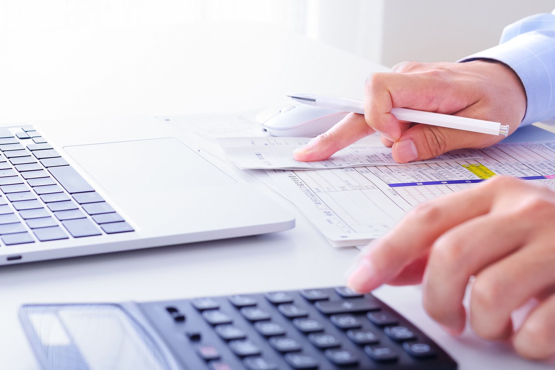 Person calculating with calculator, pointing at paperwork near laptop.