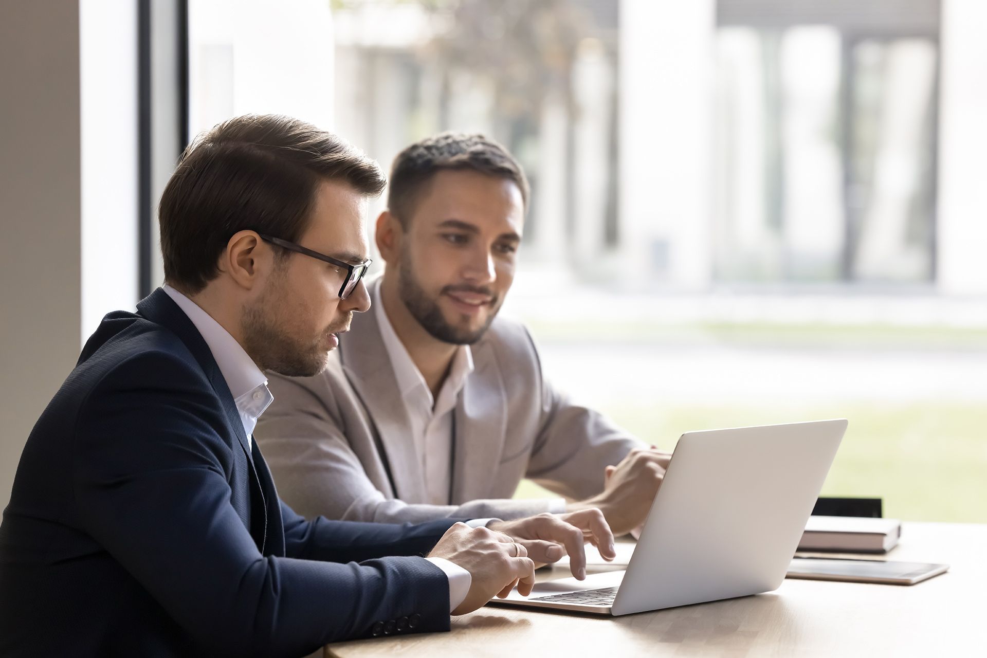 Two men in suits look at a laptop, one points, in a bright office.