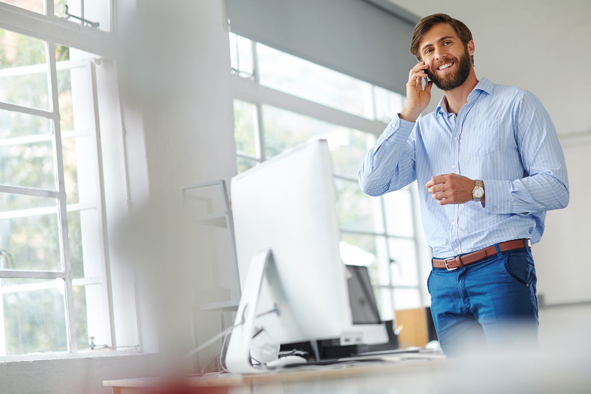Man in blue shirt, smiling, on a phone, standing near a computer and window.