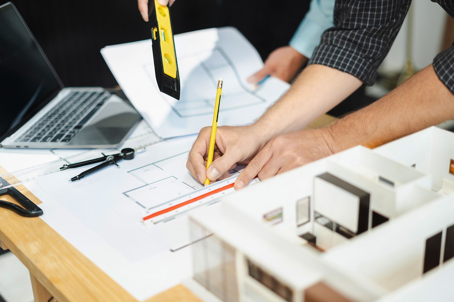 Hands sketching blueprints with ruler and pencil, alongside a model building and laptop.
