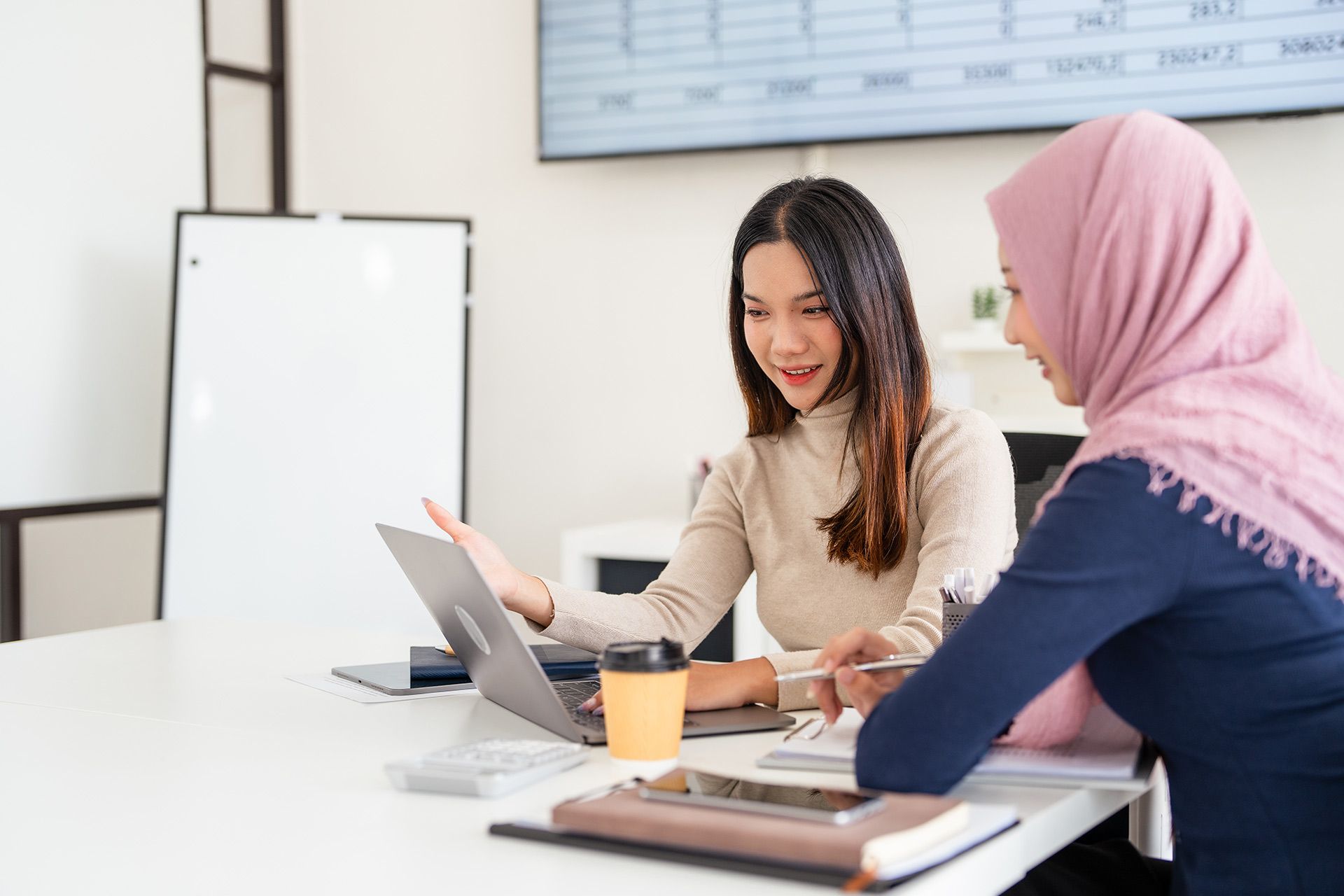 Two women collaborating at a table with a laptop, a coffee cup, and paperwork.