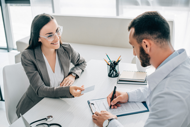 Man and woman in an office, reviewing papers. Man smiles, woman reaches to hand over a document.