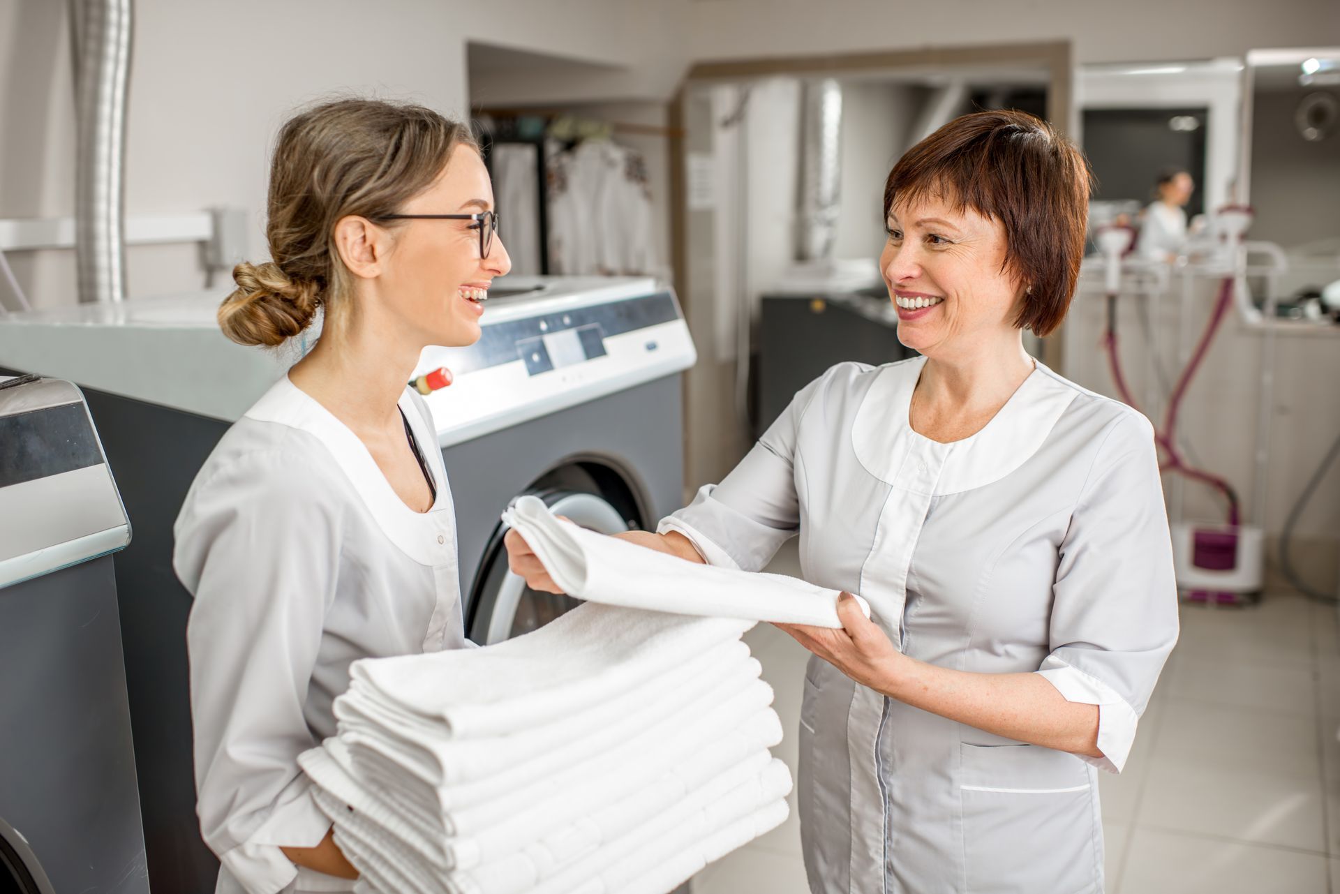 Two women are standing next to each other in a laundry room holding stacks of towels.