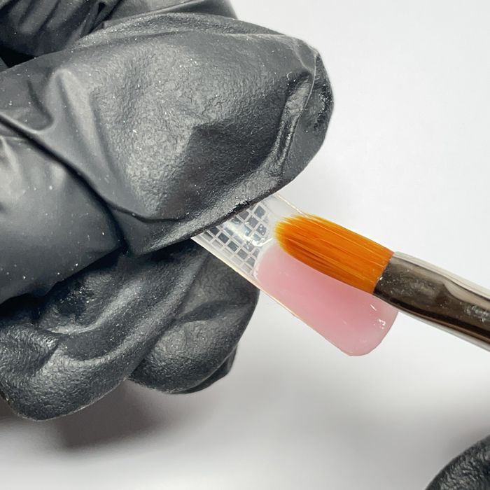 Mano guantata che usa un pennello per applicare l'acrilico rosa su una cartilagine per unghie su uno sfondo bianco.