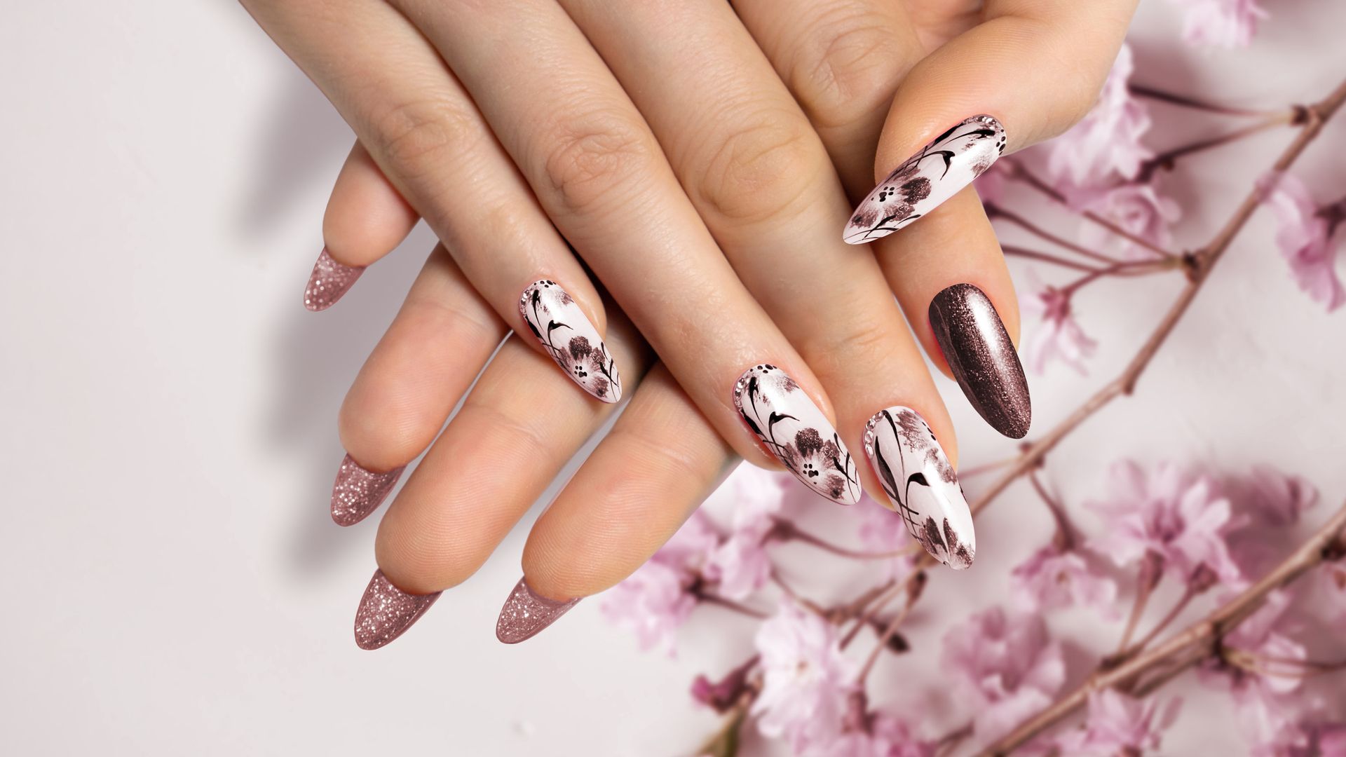 Mani con unghie lunghe a mandorla. Alcune sono bianche con motivi marroni; altre sono rosa scintillante. Fiori rosa.