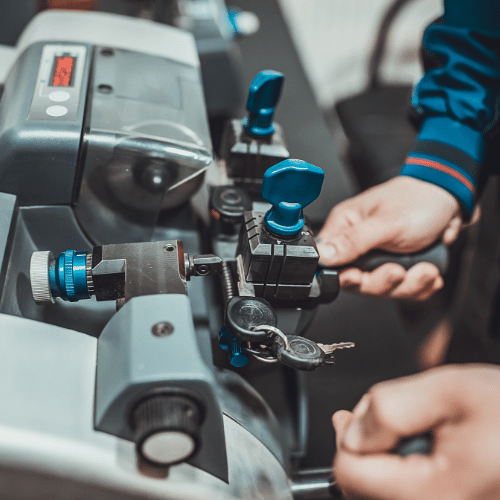 A locksmith using a key-cutting machine, with hands holding a key in place, and other keys hanging nearby.