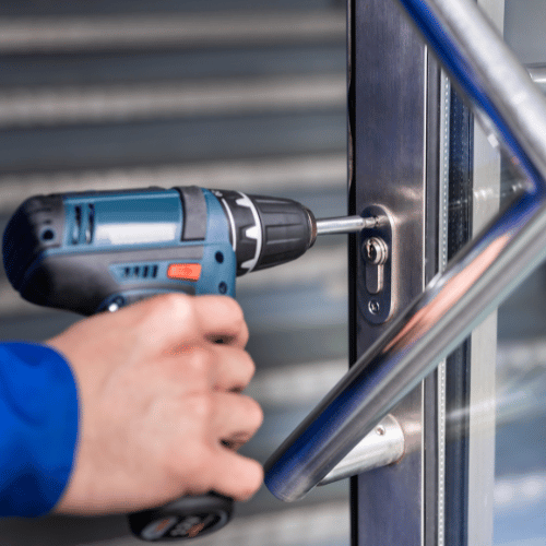 Hand using a power drill to install a lock on a metal door with a silver handle.