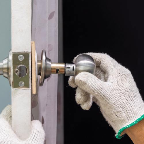 Gloved hands installing a silver doorknob on a white door frame. Black background.