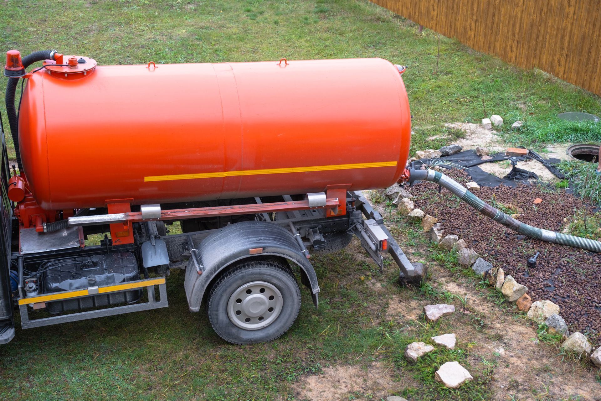 An orange water tank trailer sits on grass, with a grey suction hose connected to a patch of dark soil.