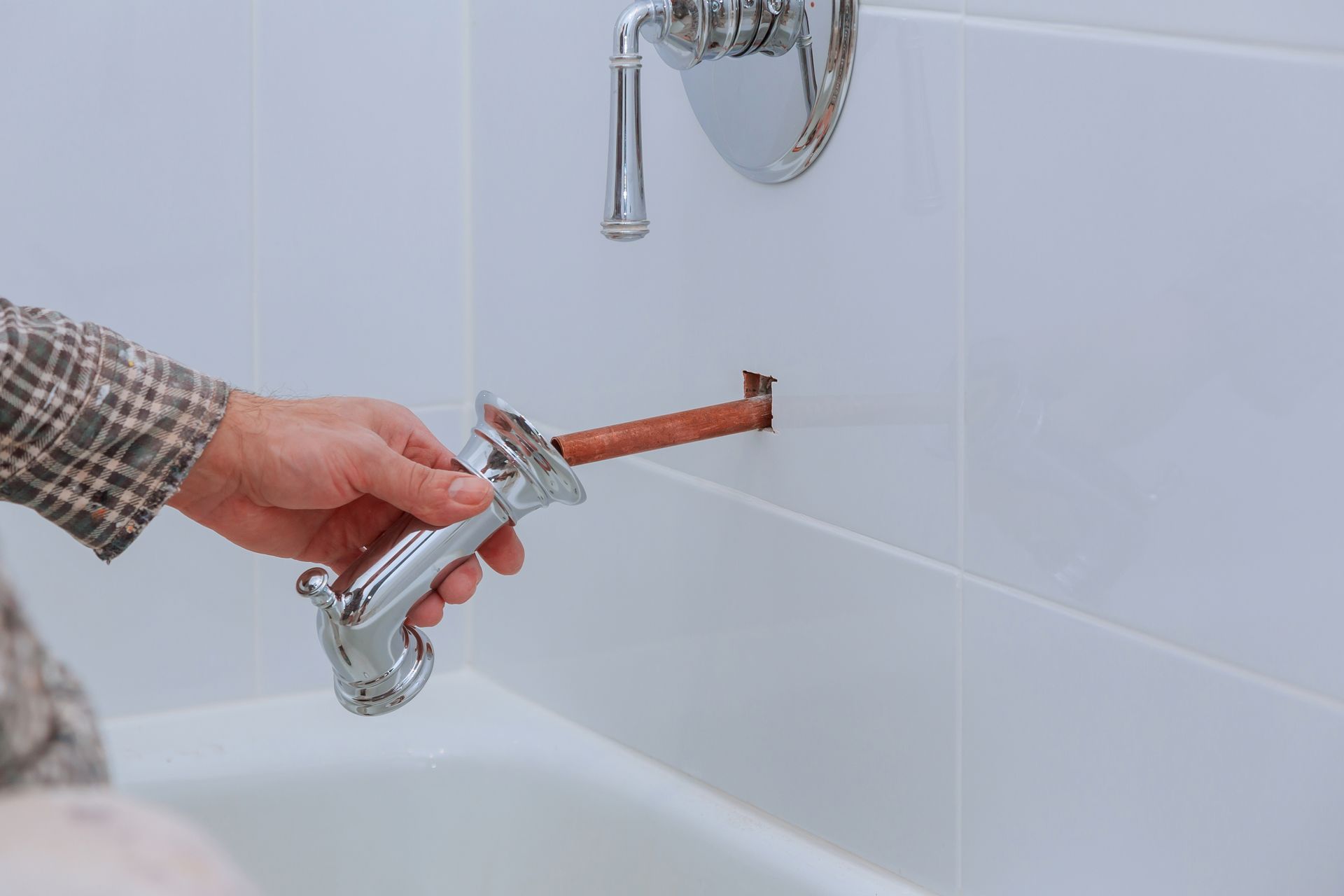 A hand holding a chrome bathtub spout in front of a copper pipe protruding from a tiled bathroom wall.