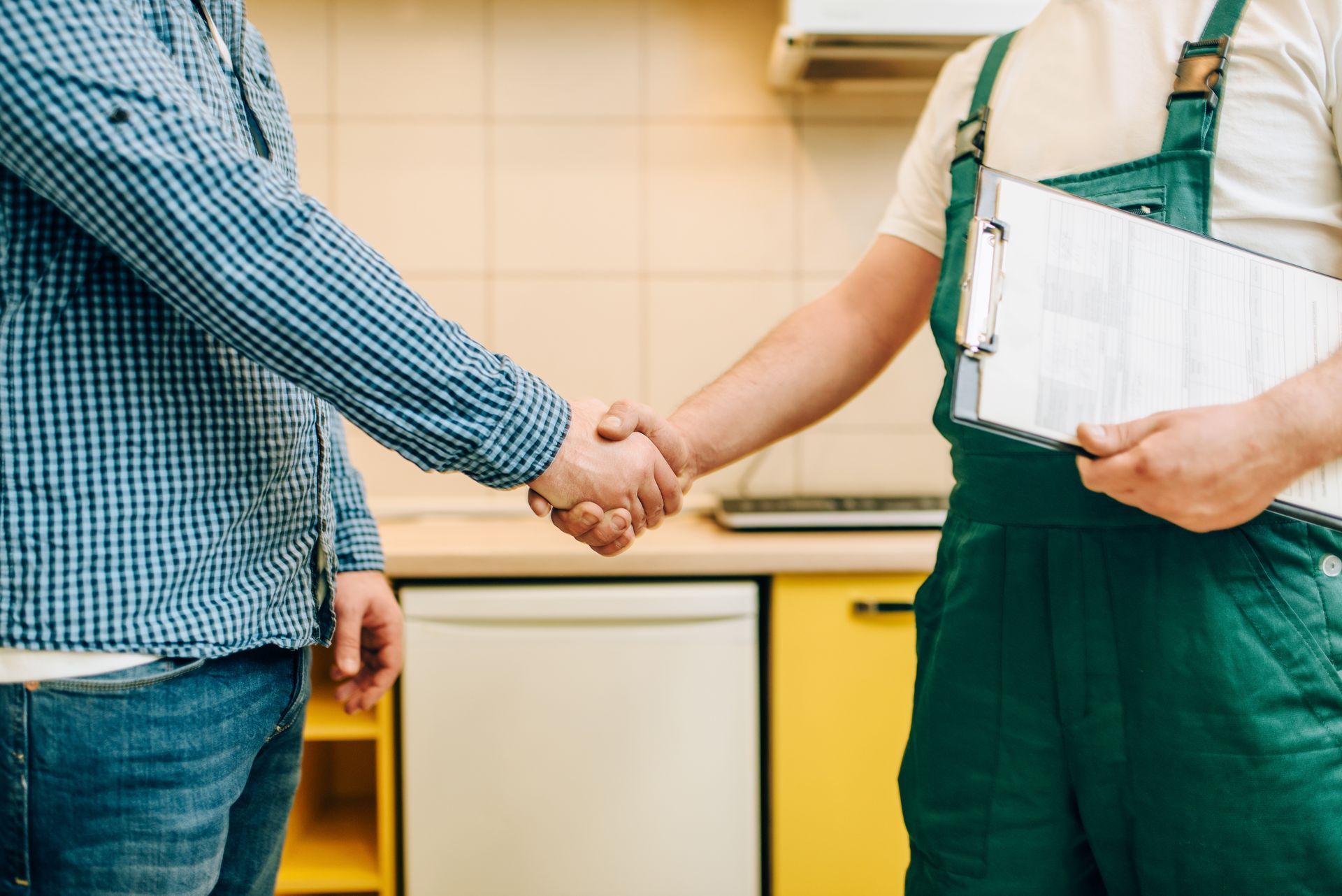 Two people shaking hands in a kitchen, one wearing green work coveralls and holding a clipboard.