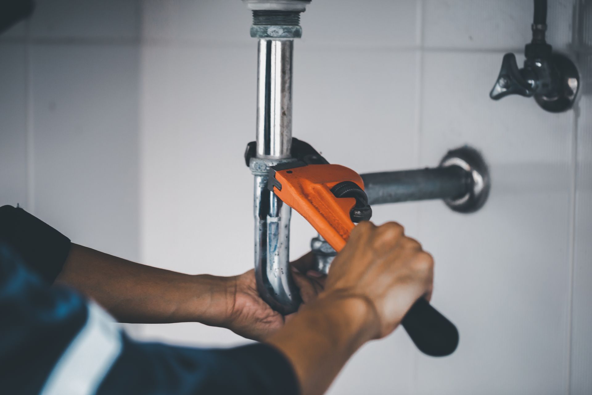 A person uses an orange pipe wrench to tighten a metal drainpipe under a sink.