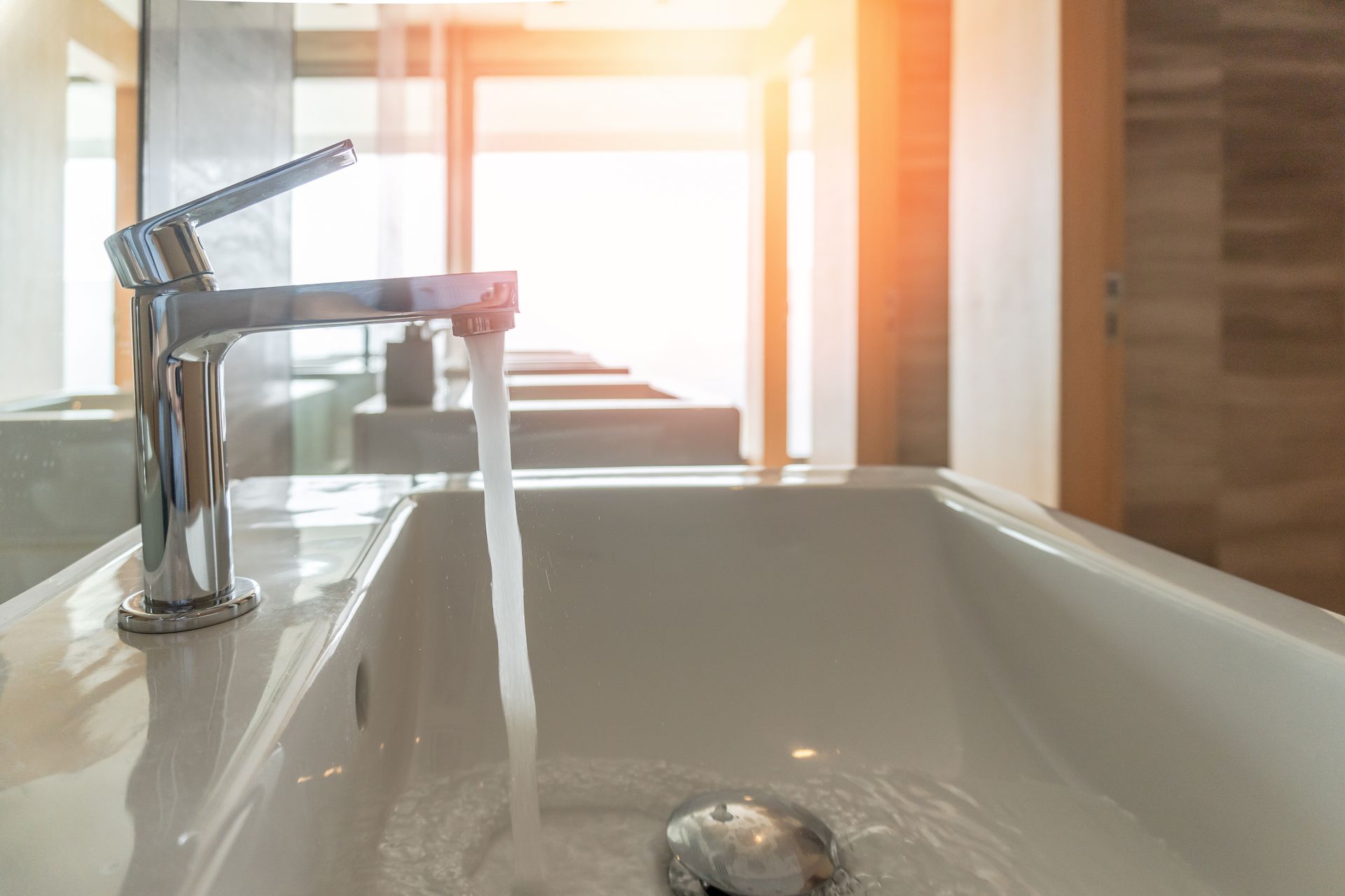 A modern chrome bathroom faucet running water into a white sink, set against a bright, sunlit room.